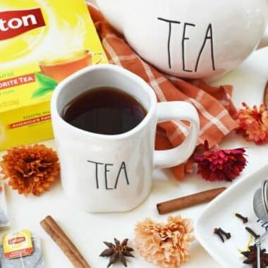 Spiced Black Tea in a mug that says tea, near a white teapot that also says tea. There are floral accents and whole spices on a white table nearby.
