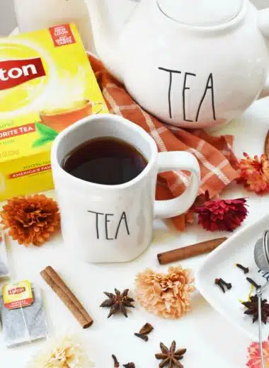 Spiced Black Tea in a mug that says tea, near a white teapot that also says tea. There are floral accents and whole spices on a white table nearby.