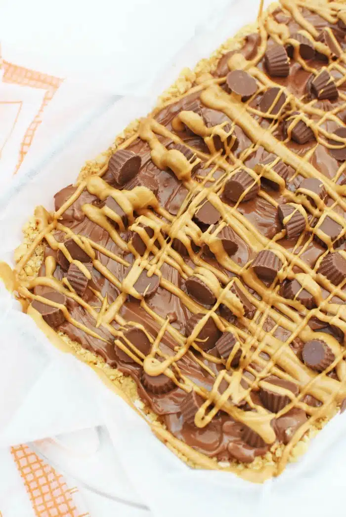 Reeses Mini Peanut Butter Cup Minis and warm melted chocolate topped onto rice crispy cereal in a pan on white table. Drizzled with peanut butter.