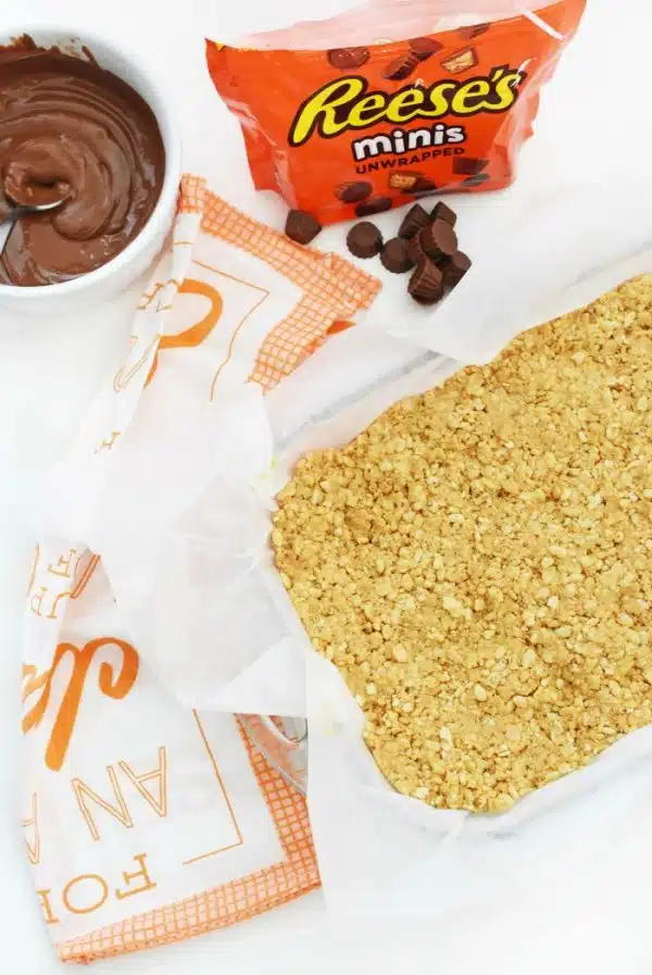 Peanut Butter Rice Cereal Bars base inside a pan lined with parchment paper on a table with reese's minis candy.