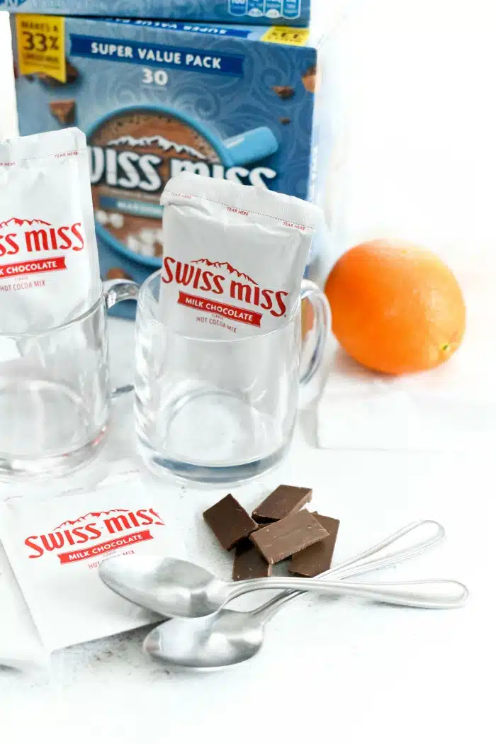 Hot cocoa packets in glass mugs with chocolate pieces and an orange on a white table. 