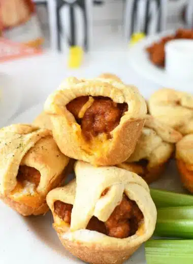 Buffalo Chicken Crescent Dough Bites on a white plate with cut celery stalks on the plate. There are black and white referee drink holders in the background.