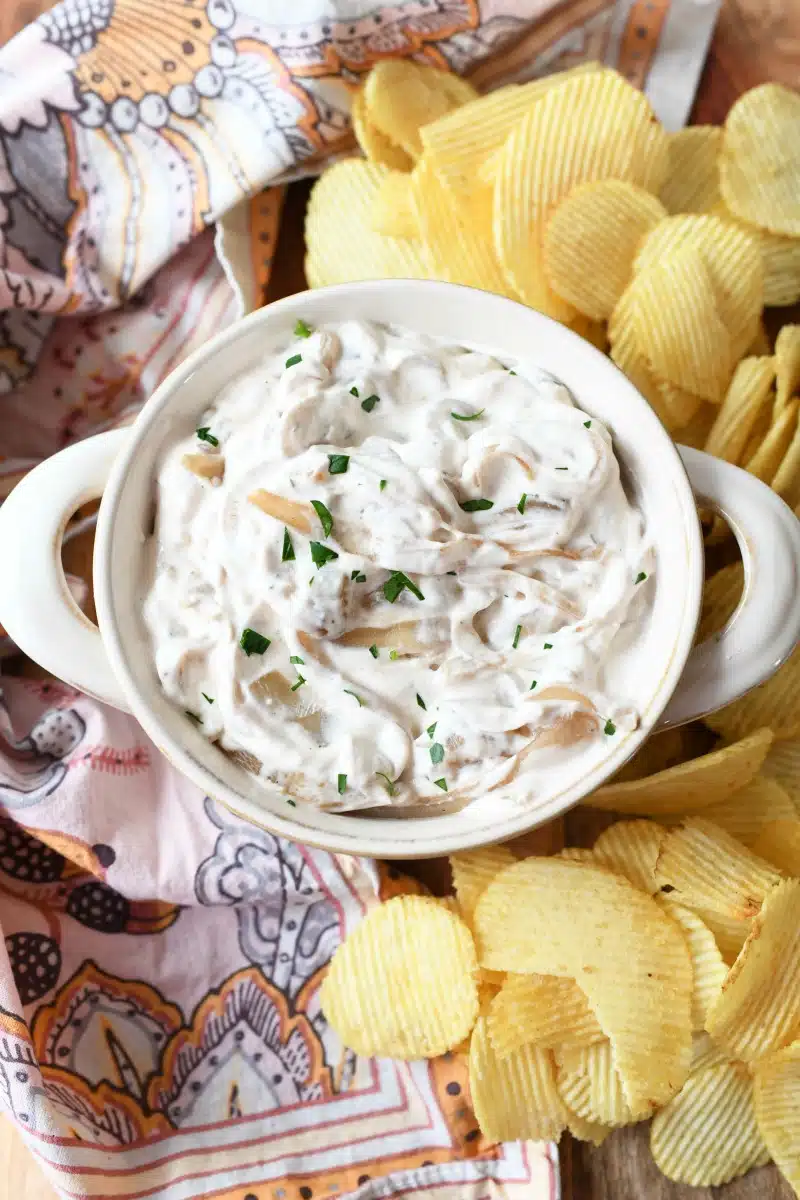 Caramelized Onion Dip on a wooden board with ruffled chips and a pink floral napkin nearby.