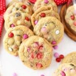 JUMBO Chewy M&M Chocolate Chip Cookies are spread on near a red dot napkin on a white table. There are 11 cookies in this image with Valentine's M&Ms scattered around.