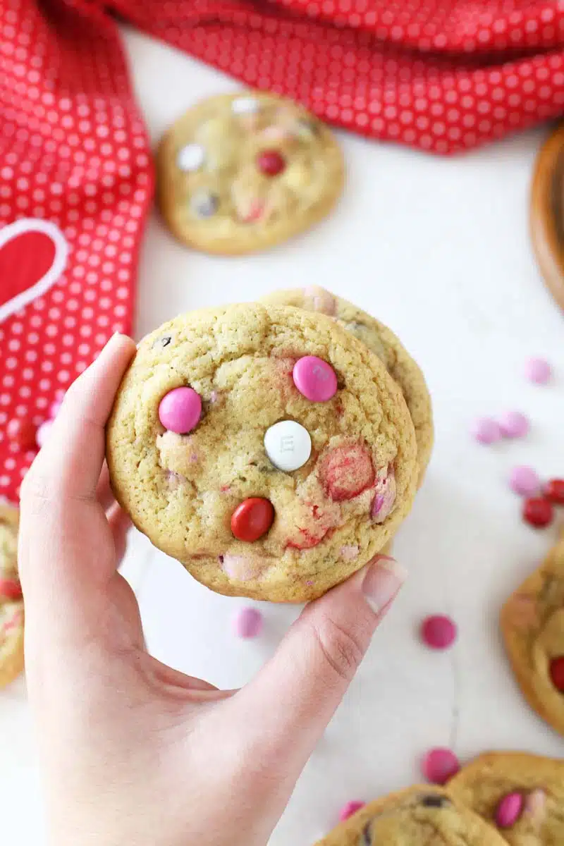 A white hand is touching a stack of Jumbo M&M Chewy Cookies. These are on a white table with a red dot napkin. Pink and red M&Ms are scattered around the table.