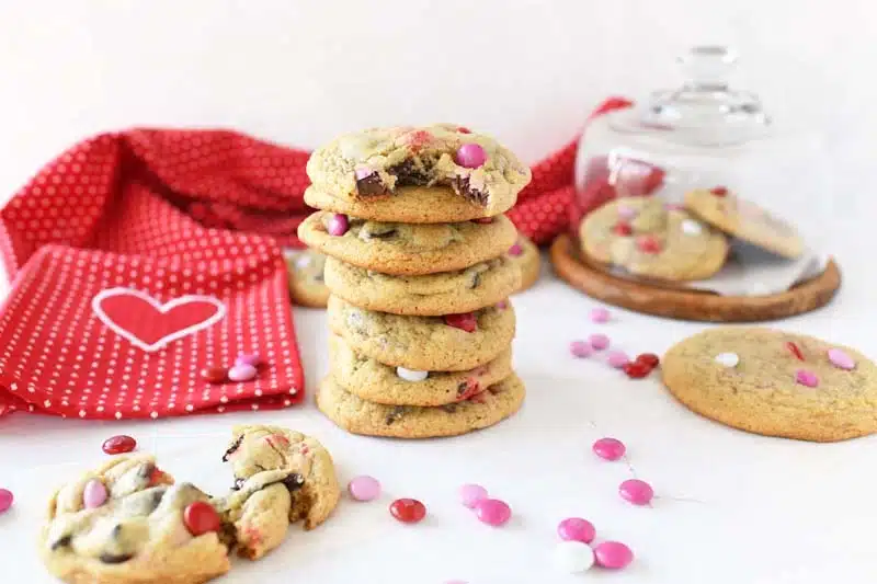 The best Valentine's Day Cookies are stacked 7 high. There is a cookie holder and red dot napkin in the image. There are pink and red M&Ms on the tabletop.
