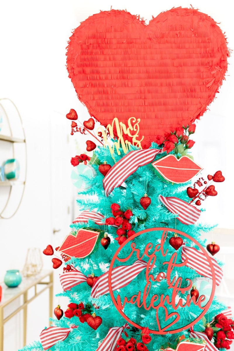 Romantic Valentines Day Tree Idea. A red and teal Valentine's Day tree with a jumbo heart pinata topper. There is red and pink décor on this tree.
