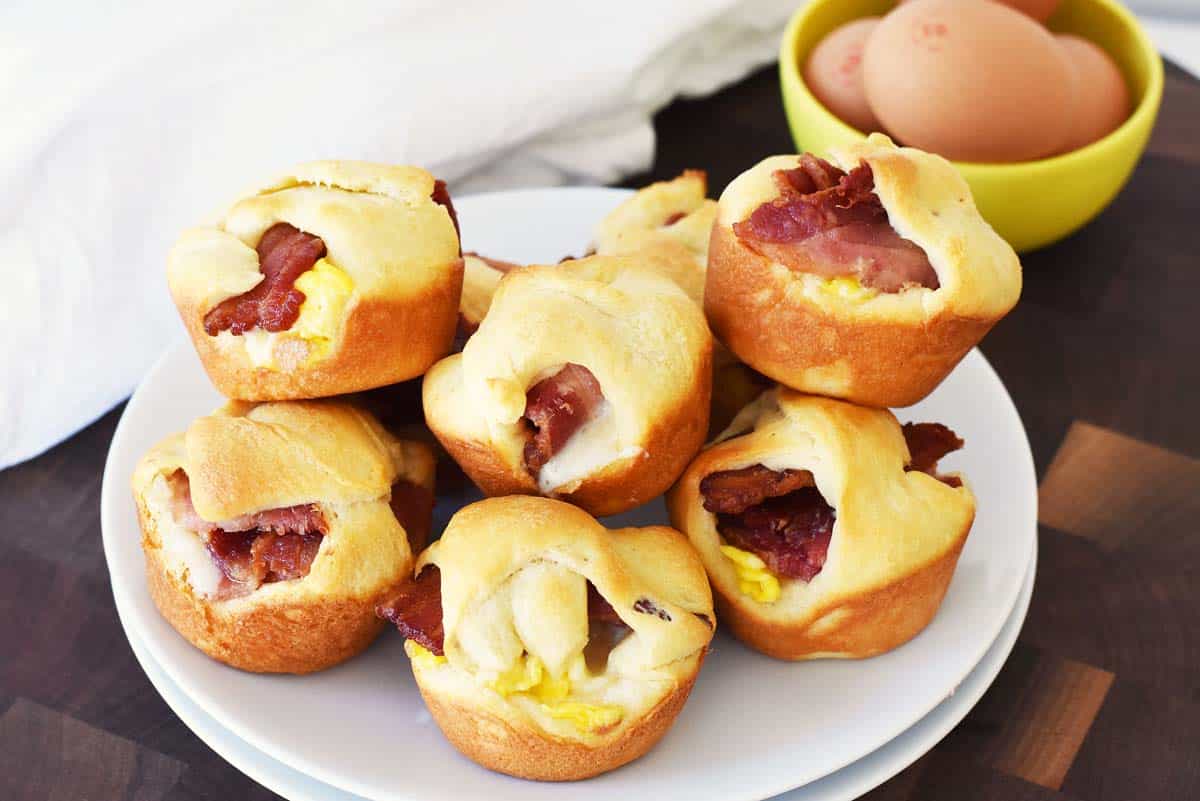Crescent Bacon and Egg Bites stacked on white plates near fresh eggs.