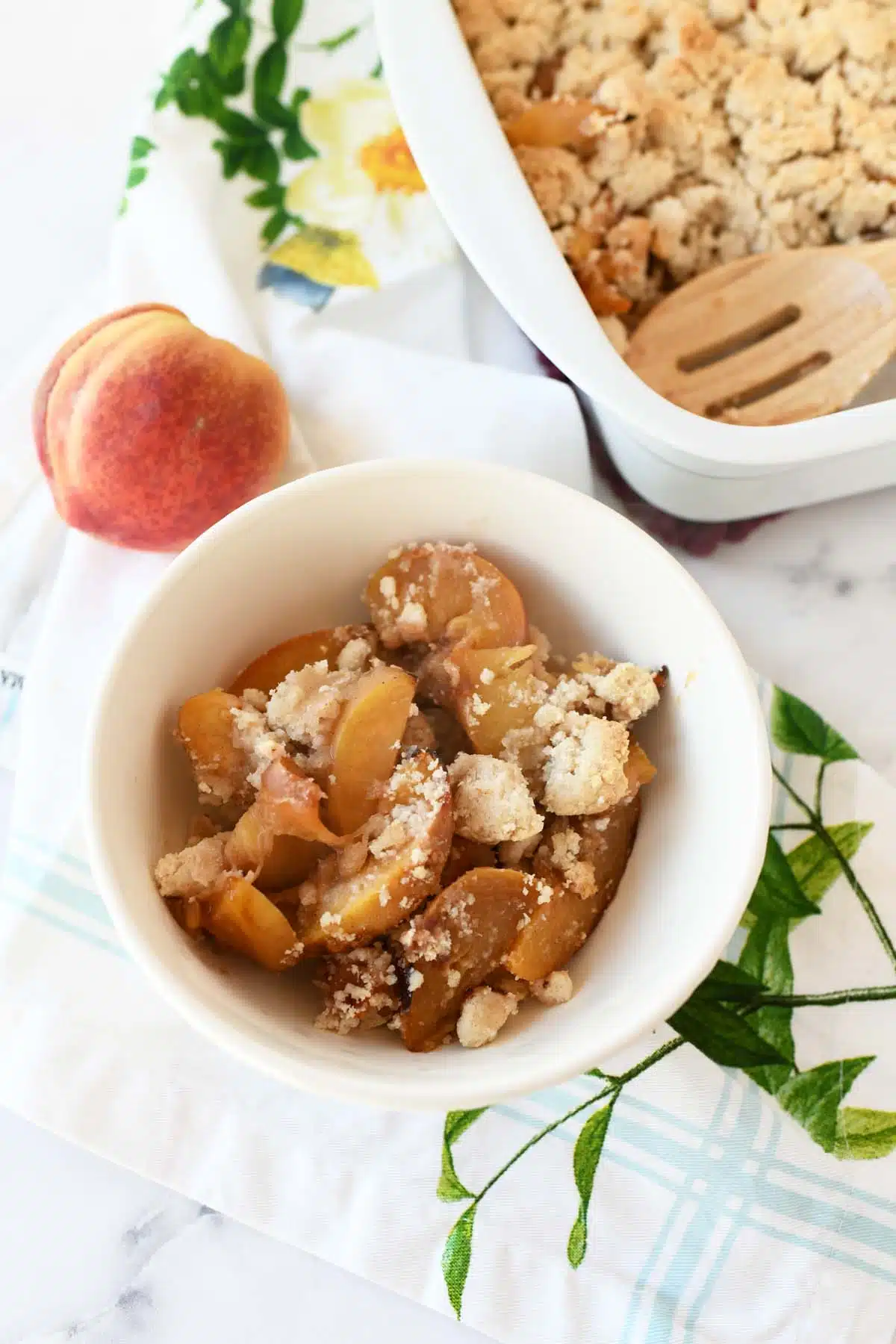Warm peach crumble in a small white bowl with a peach and floral napkin nearby. 