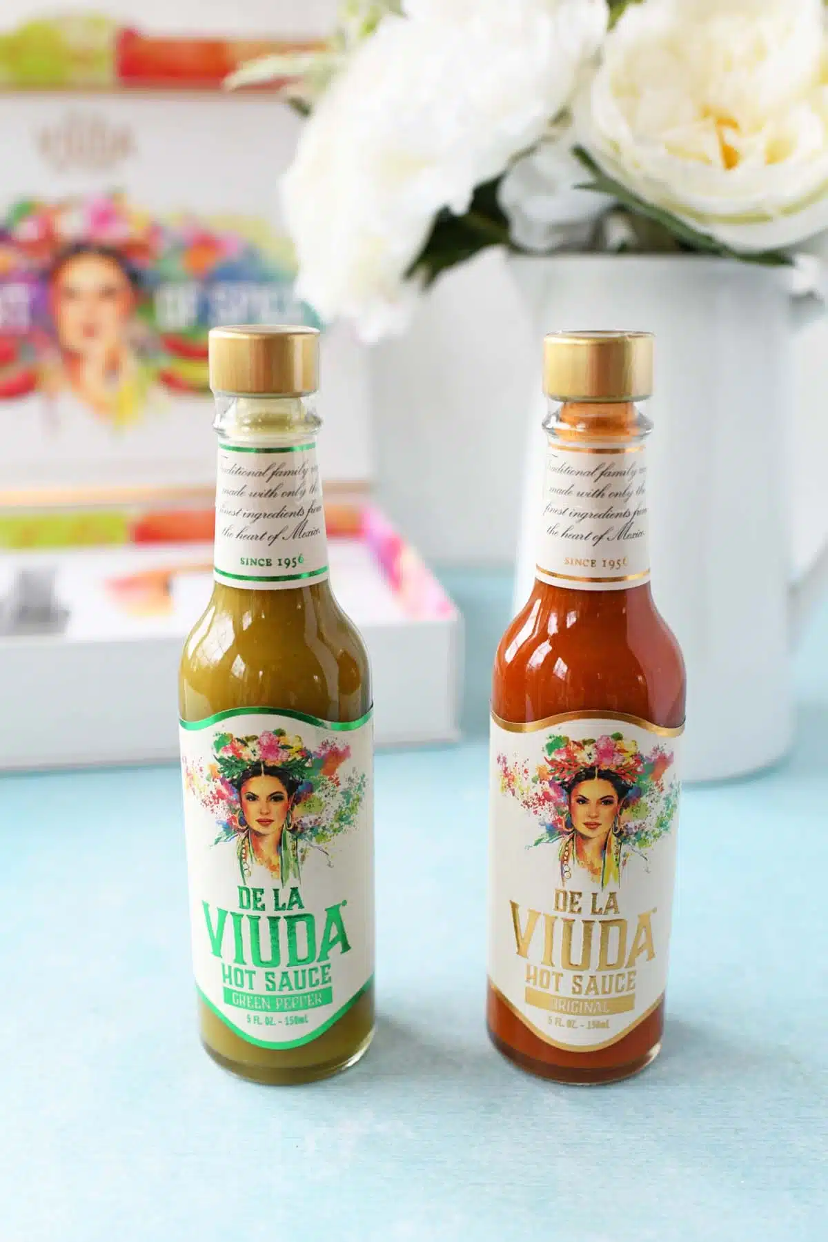 De La Viuda Hot Sauce bottles on a blue table with white flowers.