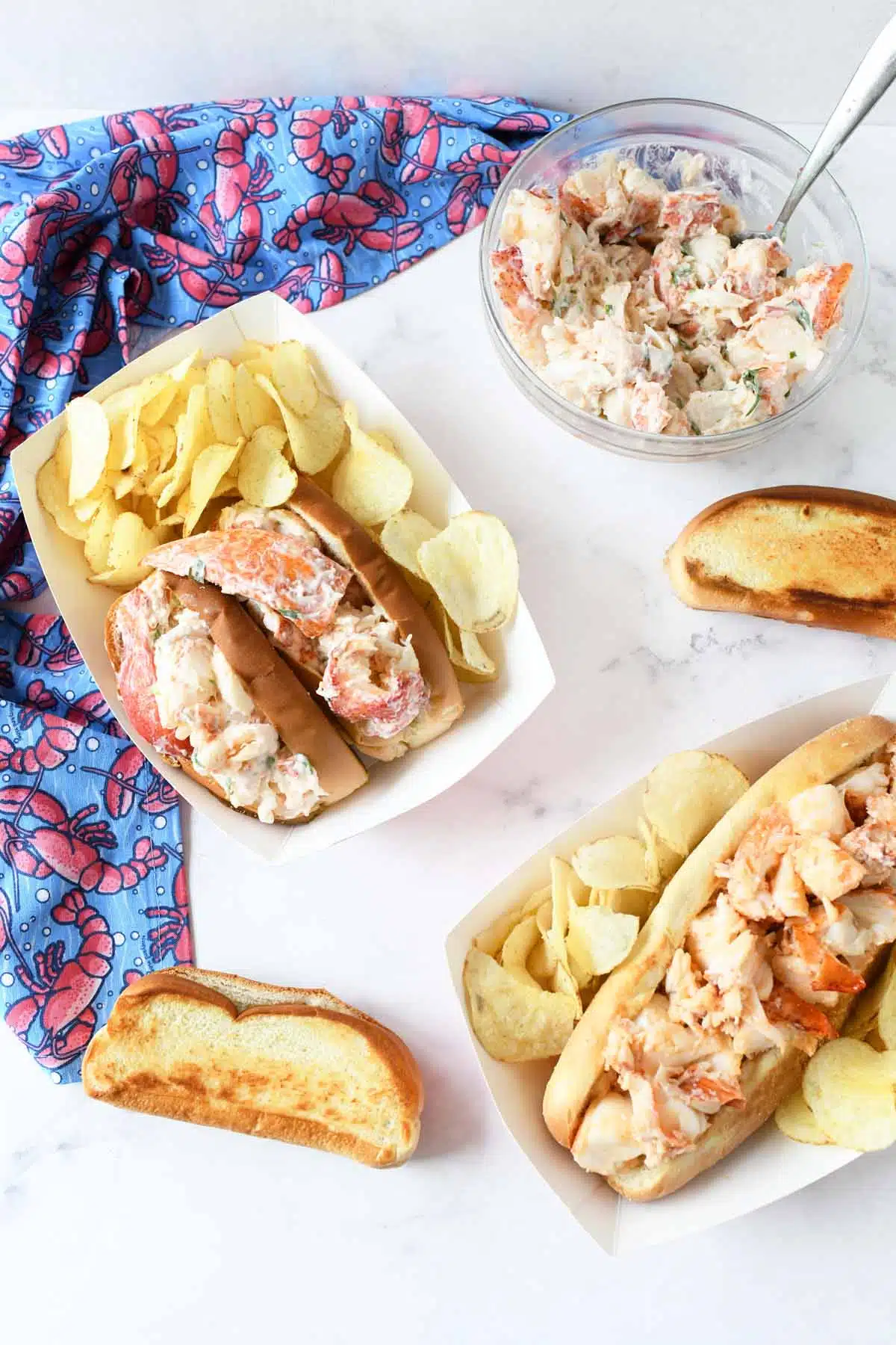 Lobster Rolls in paper containers with potato chips and a lobster napkin.
