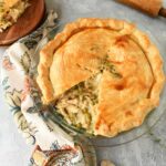 Golden chicken pot pie with a slice missing near a wooden rolling pin, and floral napkin.