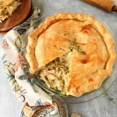 Golden chicken pot pie with a slice missing near a wooden rolling pin, and floral napkin.