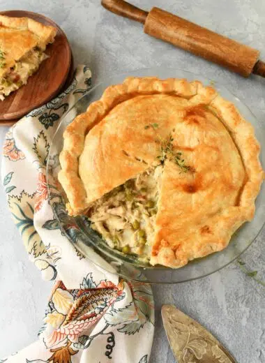 Golden chicken pot pie with a slice missing near a wooden rolling pin, and floral napkin.