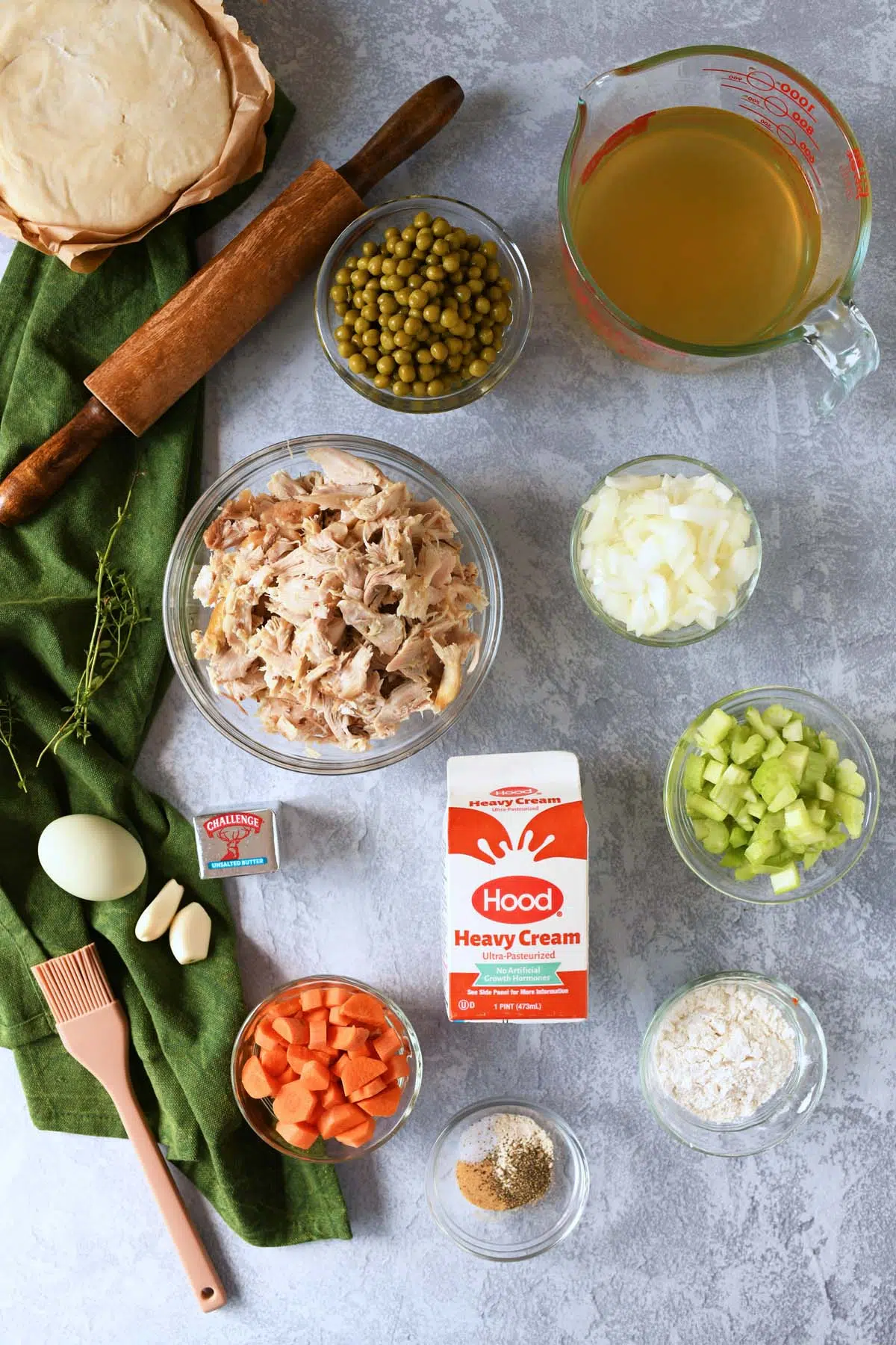 Ingredients for chicken pot pie, a rolling pin, and green napkin on a grey surface.