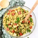 A glass bowl of colorful pesto pasta salad with a wooden spoon.