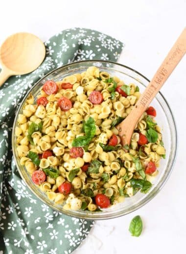 A glass bowl of colorful pesto pasta salad with a wooden spoon.