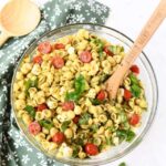 A glass bowl of colorful pesto pasta salad with a wooden spoon.