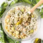 A glass bowl and spoon filled with pickle pasta salad.
