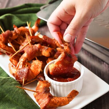 A white hand dipping smoked bacon-wrapped shrimp ina white ramekin.