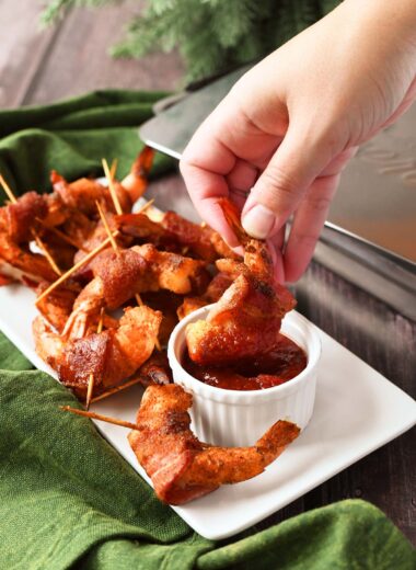 A white hand dipping smoked bacon-wrapped shrimp ina white ramekin.