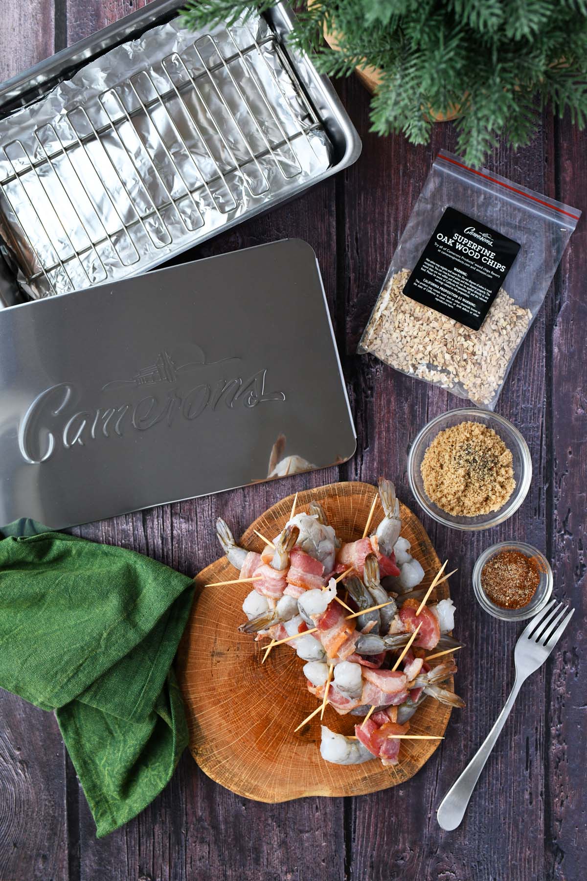 A silver smoker, and raw bacon-wrapped shrimp on a wooden table with a green napkin.