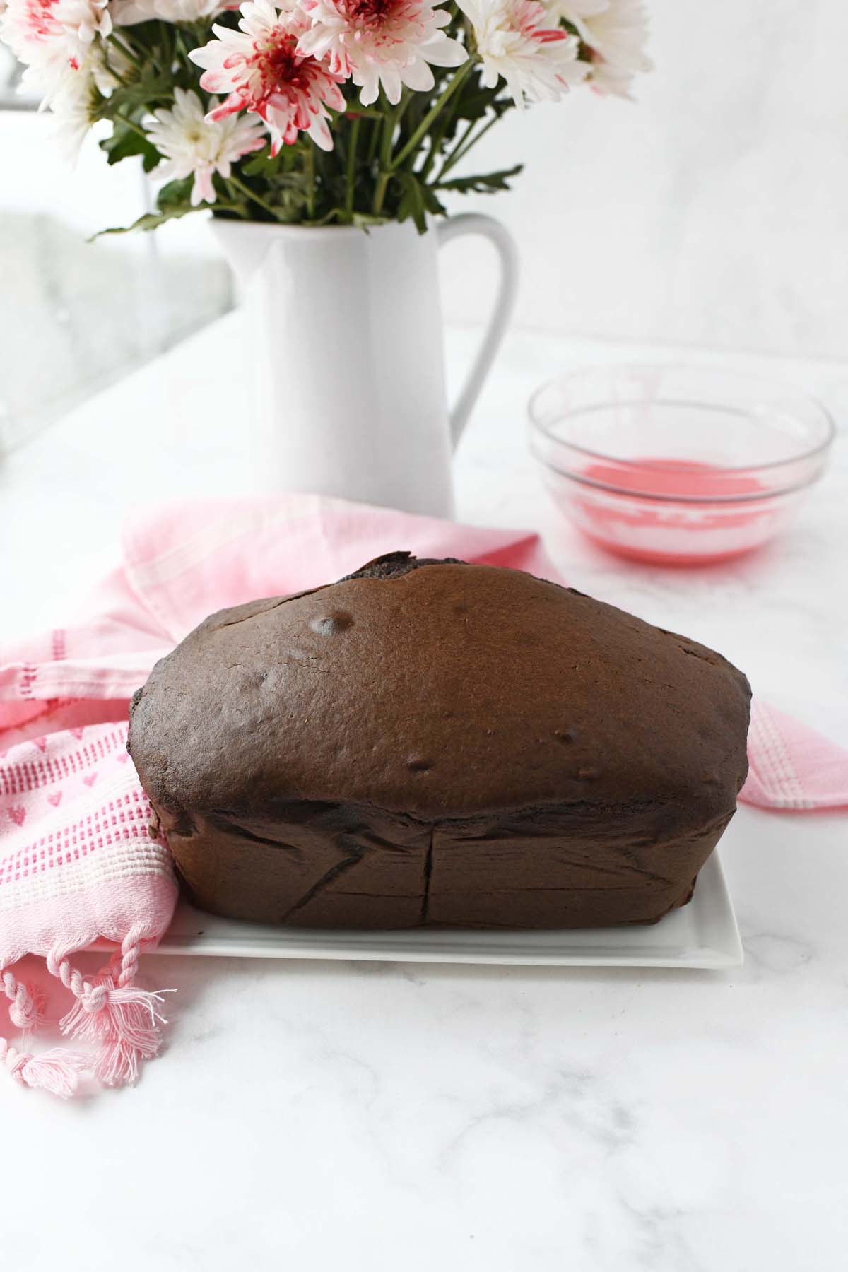 A fluffy, baked chocolate loaf cake near fresh flowers and a pink napkin.