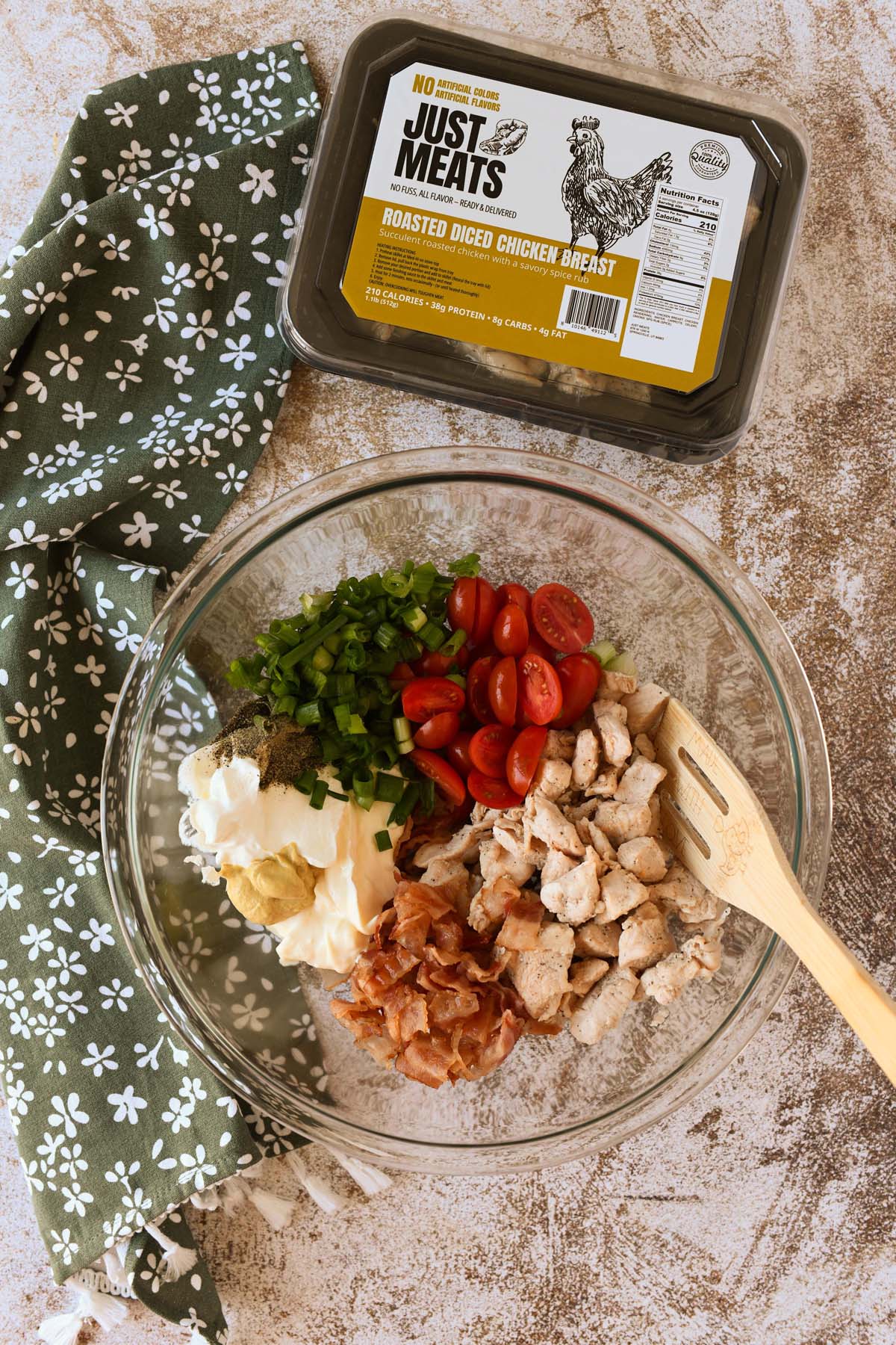 A glass bowl with BLT chicken salad ingredients inside. A wooden spoon and gren napkin is nearby.