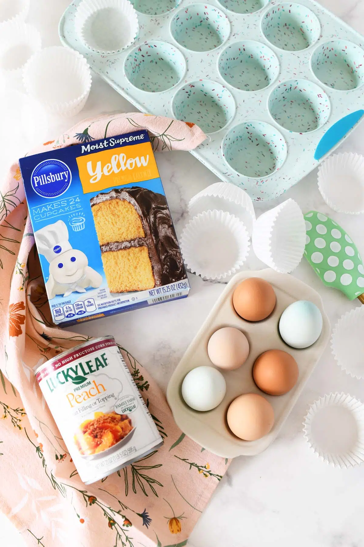 Cake mix, peach pie filling, eggs, a pink floral napkin and blue muffin tin on a white table.