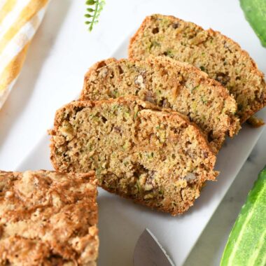 Golden-brown slices of cucumber bread on a white table with cucumbers nearby.