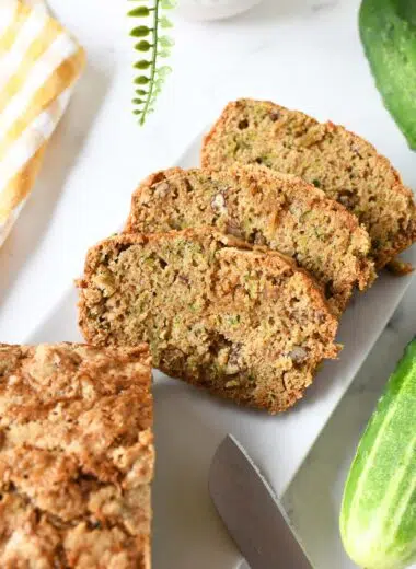 Golden-brown slices of cucumber bread on a white table with cucumbers nearby.