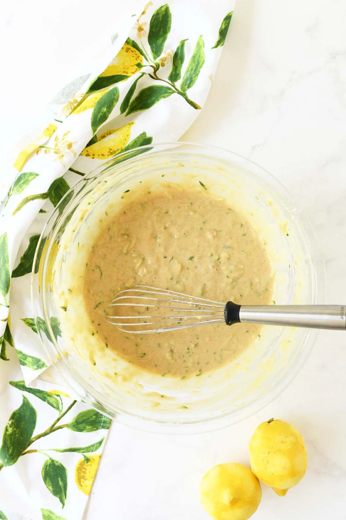 Zucchini batter in a glass bowl with a whisk and fresh lemons and a lemon napkin,