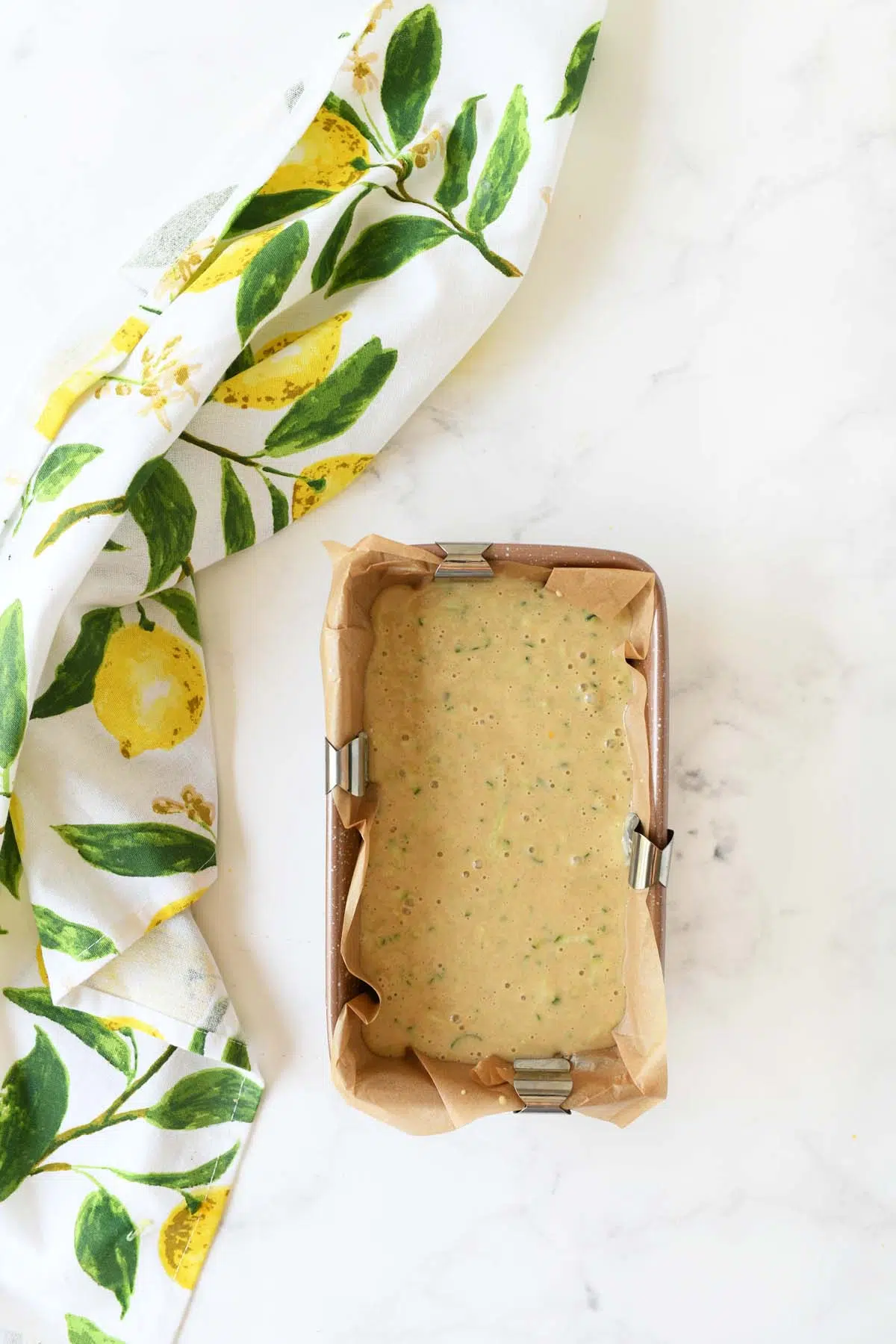 Zucchini batter in a loaf pan with clips near a lemon napkin.