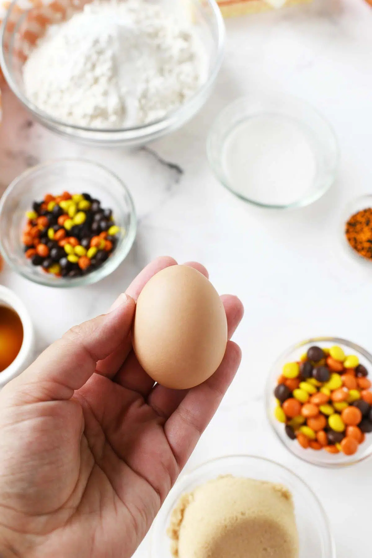 A white hand holding a brown egg with dishes of candy in the background.