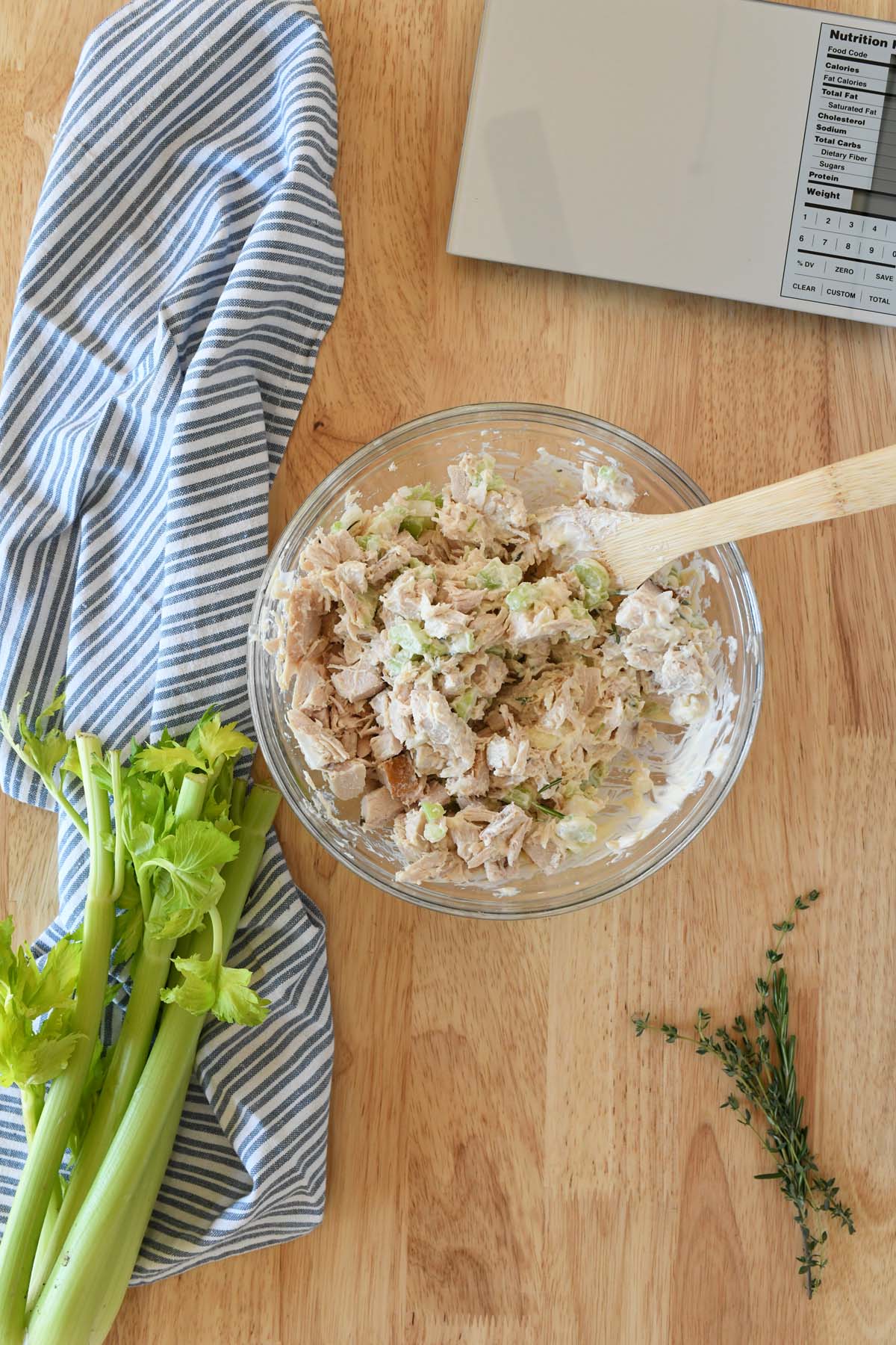 A glass bowl with turkey salad inside and a wooden spoon. There is also a blue, striped napkin and green celery nearby.