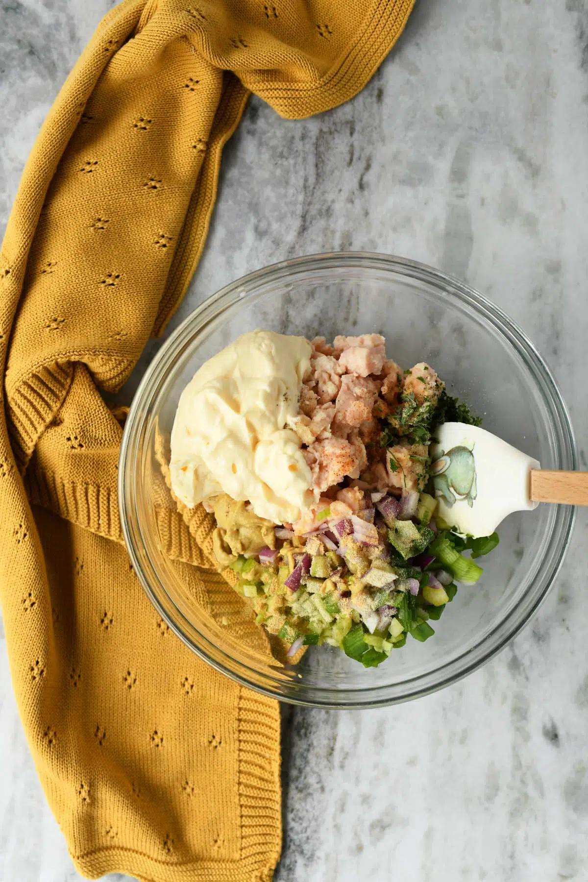Chicken Salad ingredients in a glass bowl with a white spatula and a yellow napkin.