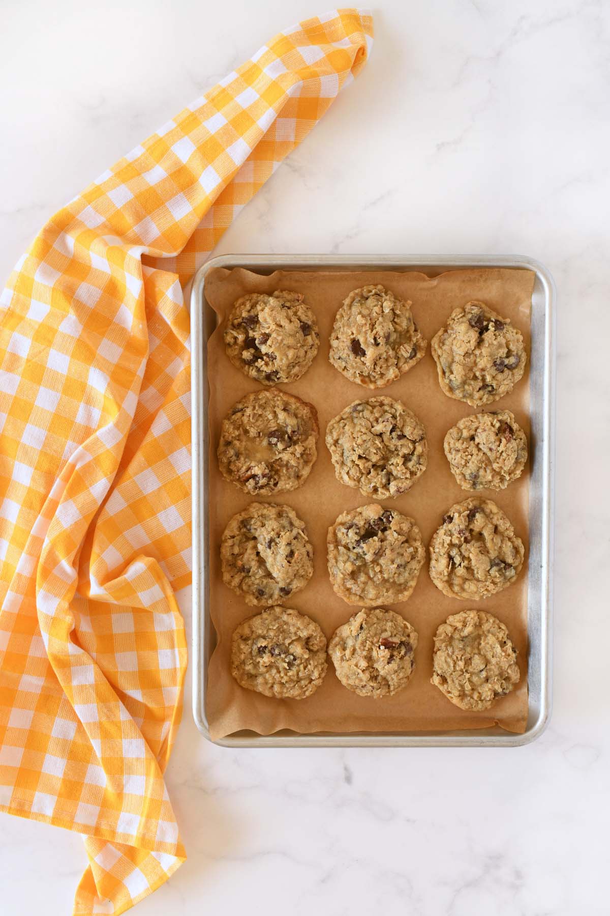 Baked cowboy cookies on a natural parchment line baking sheet near a yellow, checkered napkin.