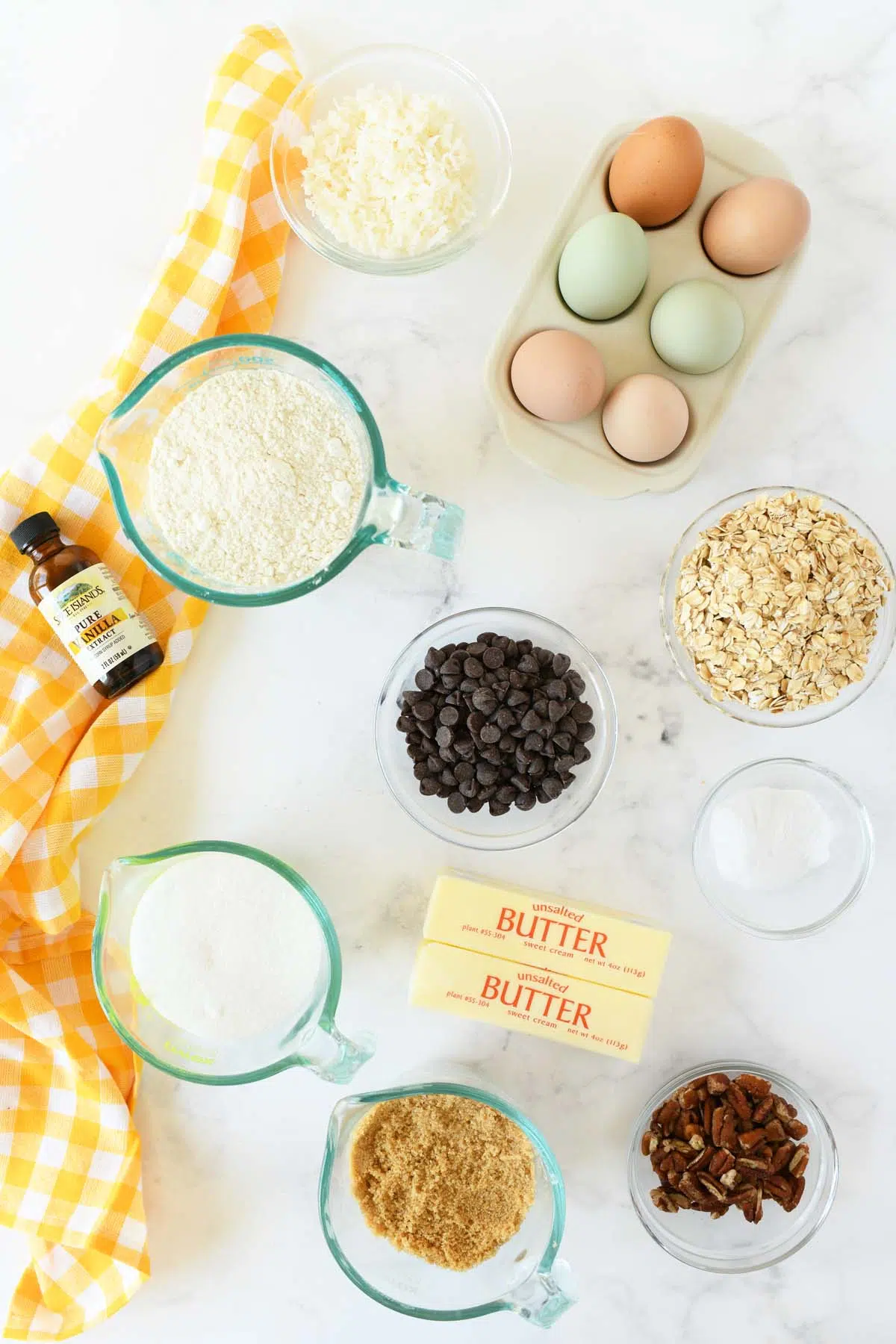 cowboy cookies ingredients in glass bowls on a white table with a yellow, checkered napkin nearby.