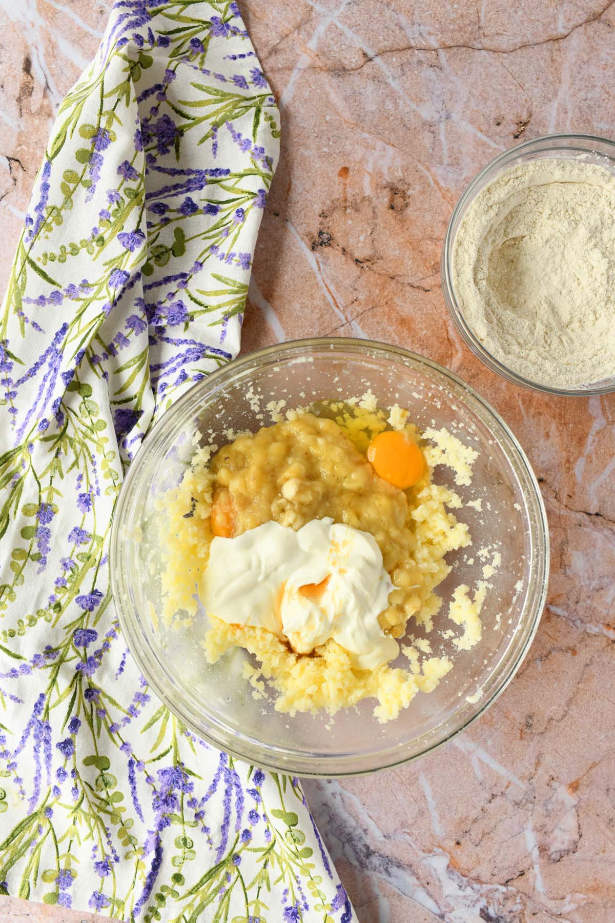 Banana bread batter in a glass bowl near a purple, floral napkin.