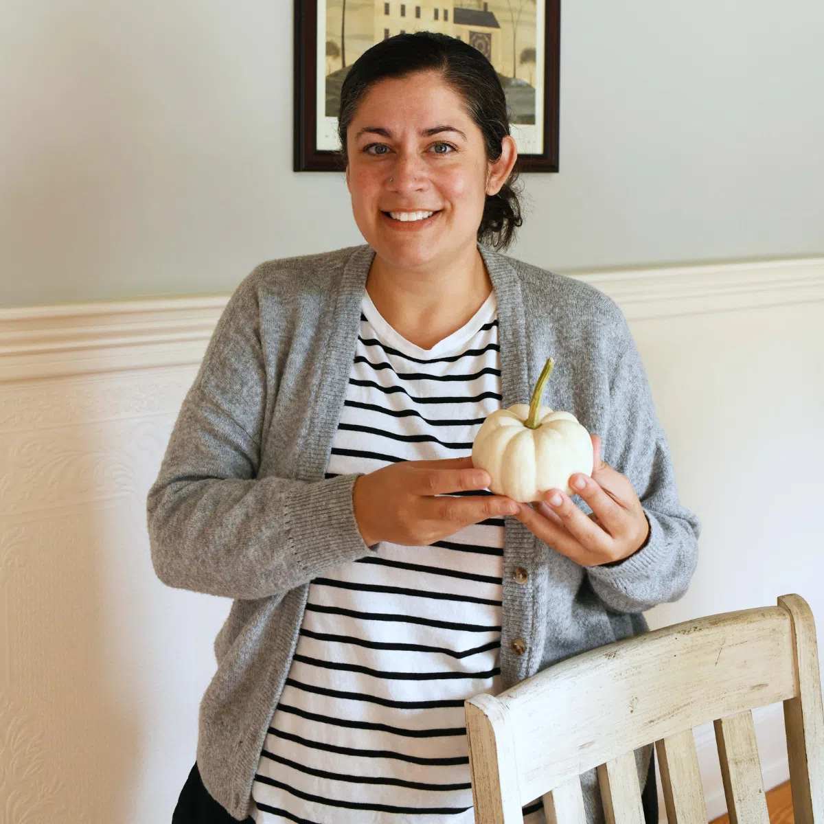 A smiling woman in a grey sweater holding a white, mini pumpkin.