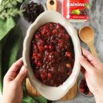 Hands holding up a scalloped grey dish of orange cranberry sauce with raisins.