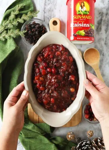 Hands holding up a scalloped grey dish of orange cranberry sauce with raisins.