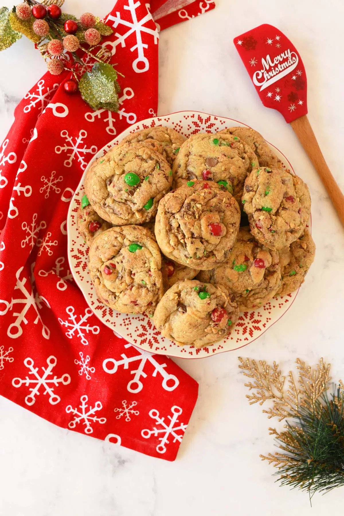 Holiday themed M&M cookies on a plate near a red, and white snowflake napkin and a red holiday spatula.