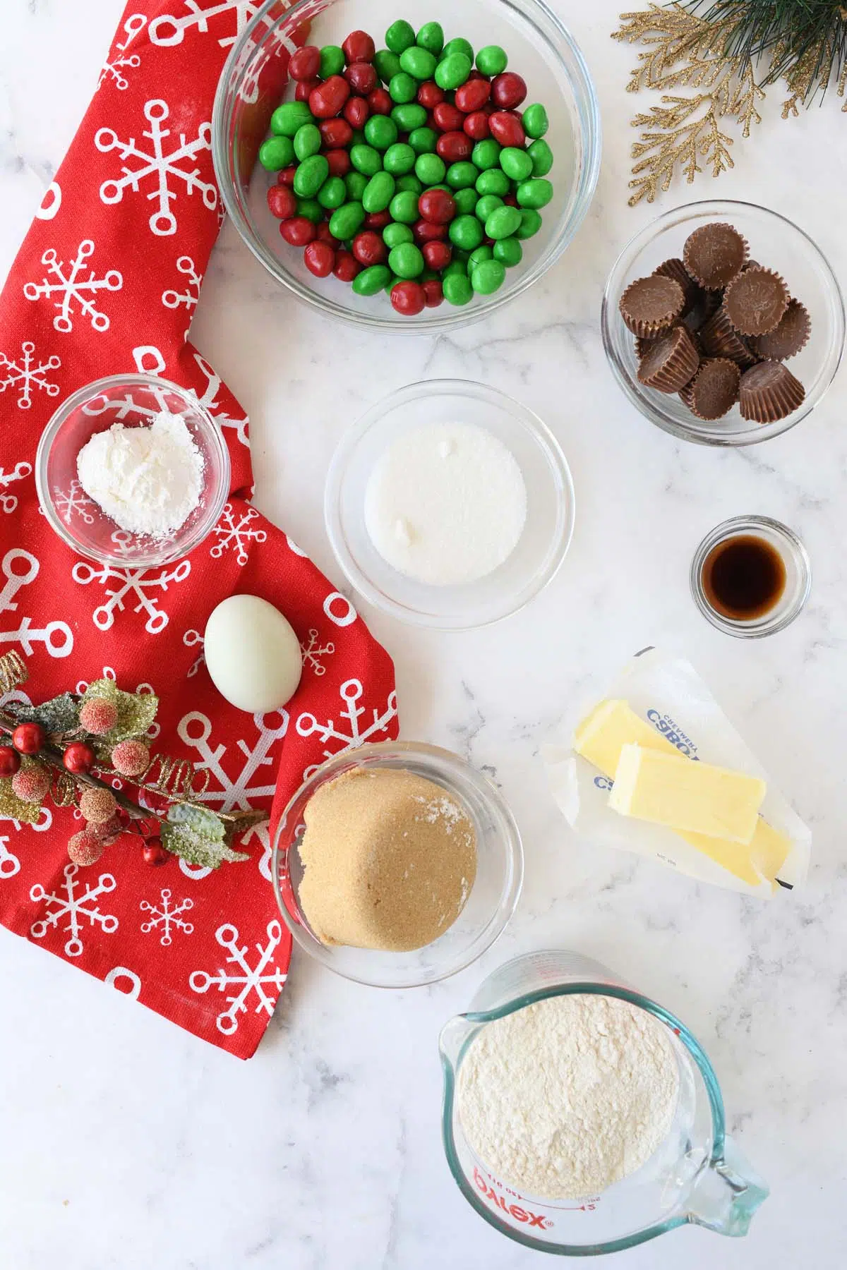 Holiday M&M and peanut butter cup ingredients in glass bowls near a red, snowflake napkins.