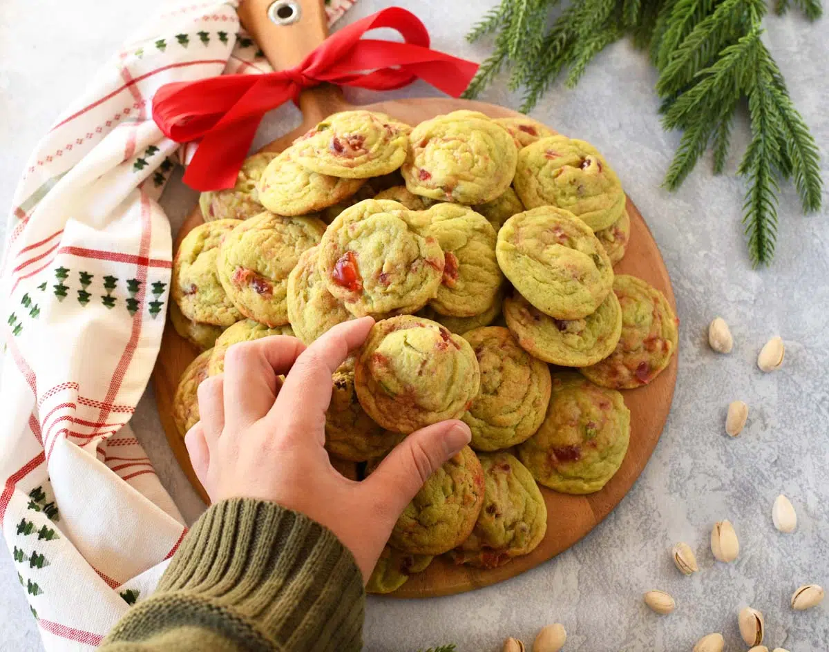 A hand grabbing a pistachip cherry cookie off a round, wooden board with a red bow.
