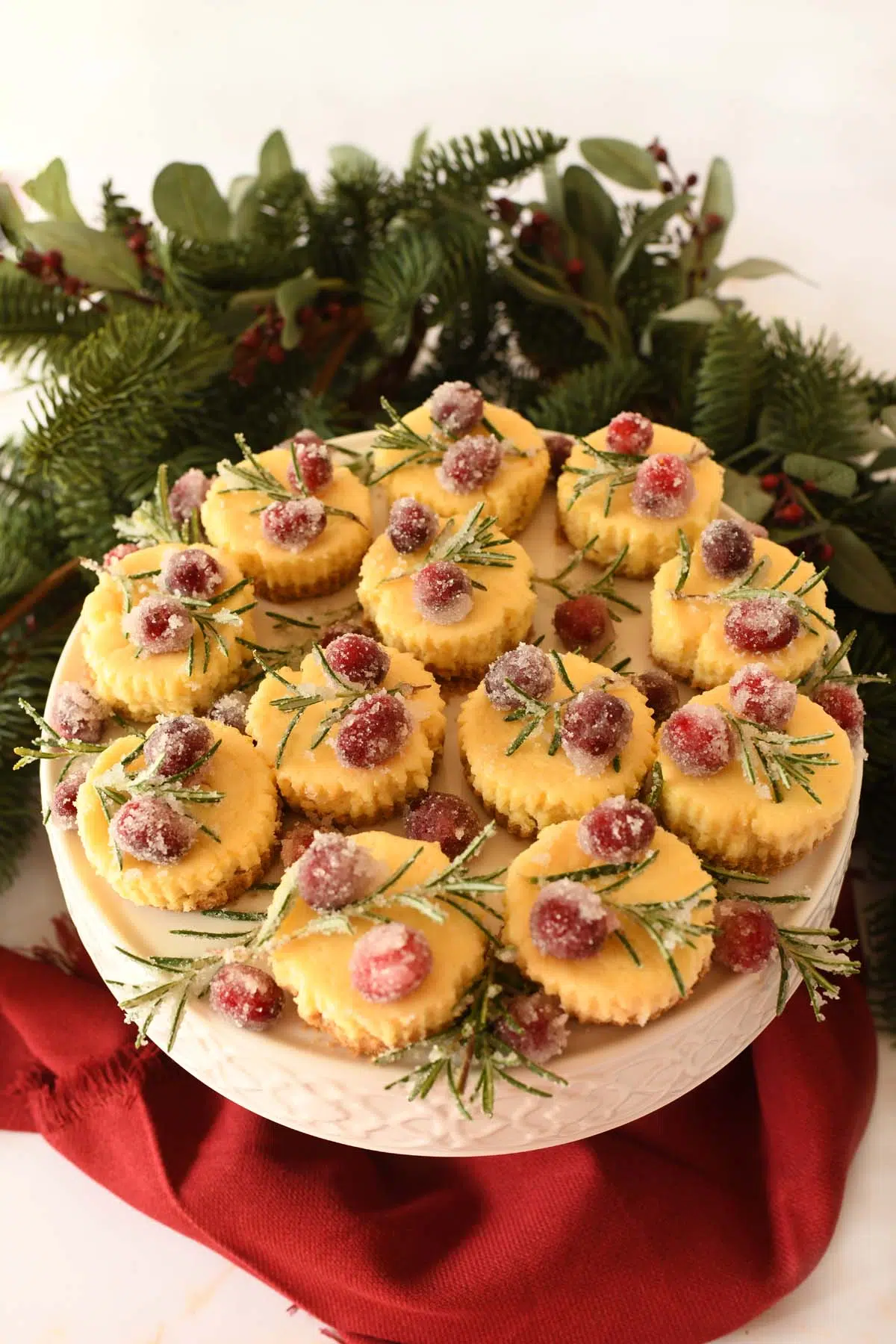 Festive mini eggnog cheesecakes on a stand with candied cranberry and rosemary on top, near a red napkin.