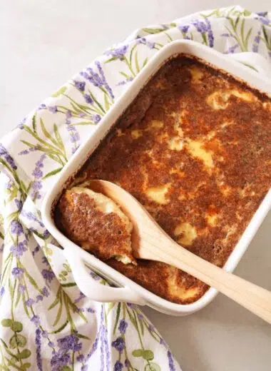 A white dish of Grape-Nuts Pudding with a wooden spoon inside. A printed purple floral napkin is also nearby.