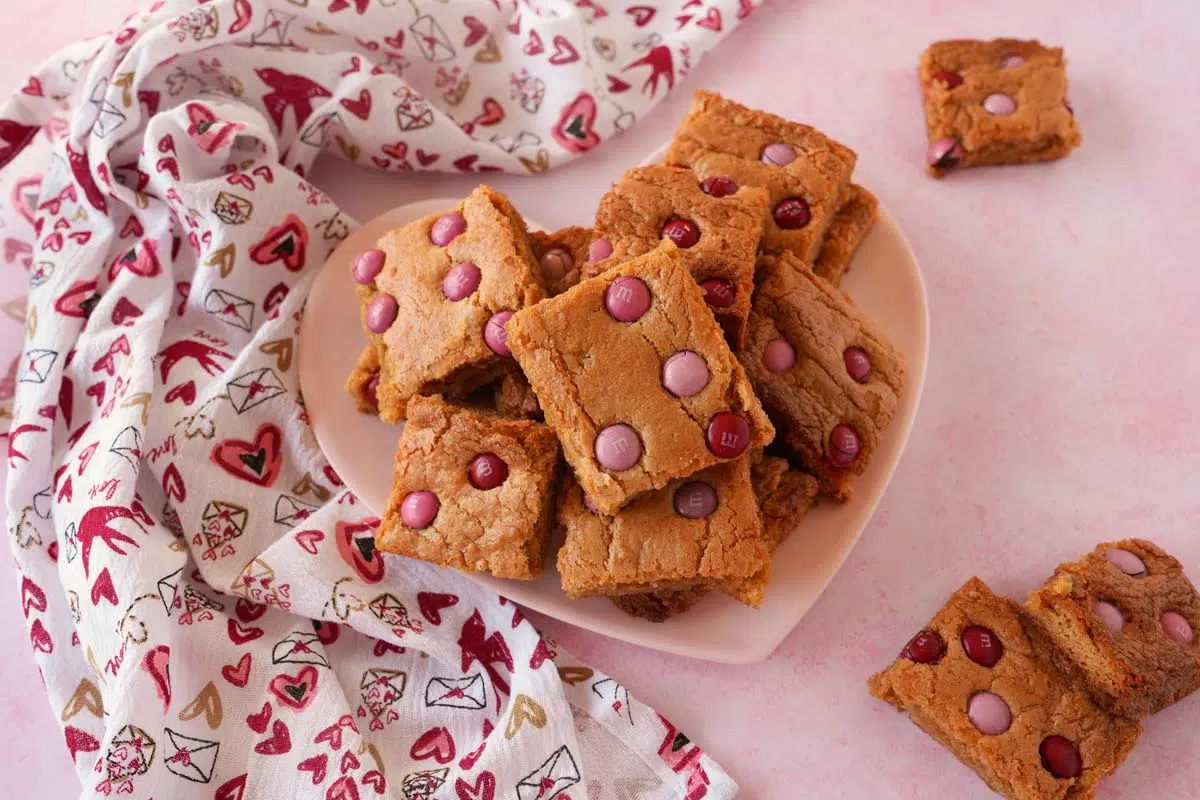 A pink heart plate is stacked with festive peanut butter blondies on a pink table, near a festive napkin.