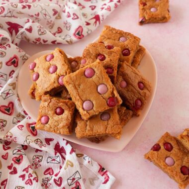 A festive plate of blondies in nestled on a pink, heart plate near a festive napkin on a pink table.