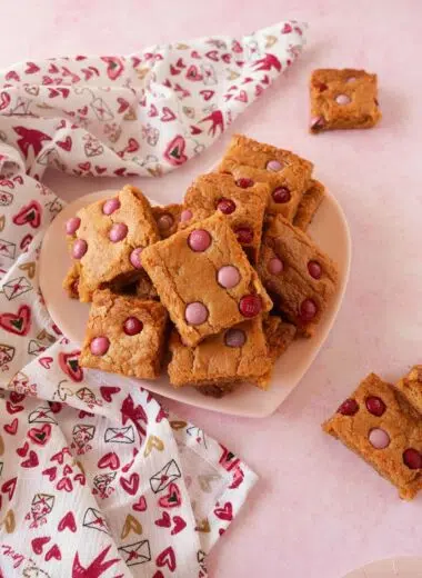 A festive plate of blondies in nestled on a pink, heart plate near a festive napkin on a pink table.